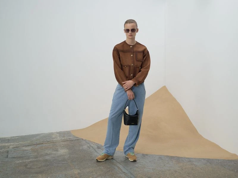 Adult poses in a brown jacket, light blue pants, and sunglasses, holding a black purse, standing indoors near a geometric design.