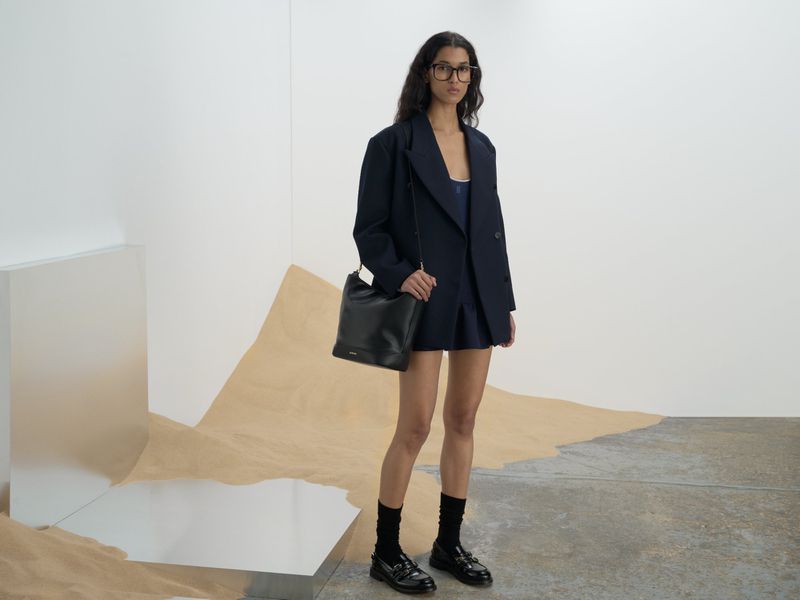 A woman stands in a studio, modeling a navy oversized blazer and matching skirt, paired with black socks, and shiny loafers.