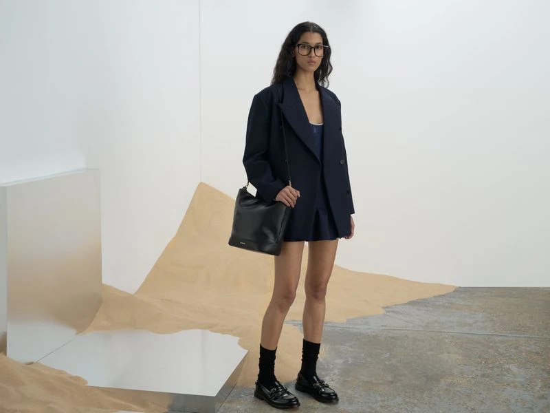 A woman stands in a studio, modeling a navy oversized blazer and matching skirt, paired with black socks, and shiny loafers.