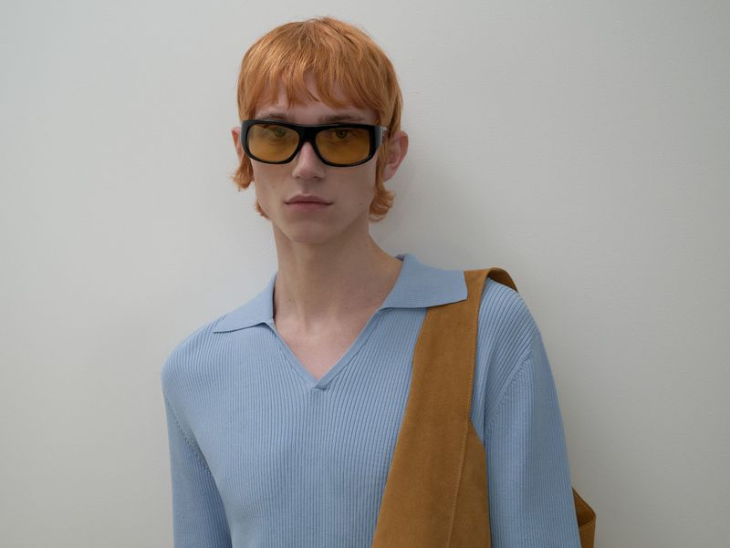 Man with orange hair wears a blue long-sleeve shirt, yellow tinted glasses, and tan shoulder bag against a neutral background.
