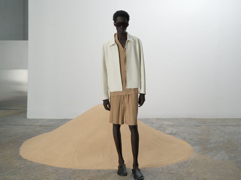 A slender man in a cream jacket and matching sand-colored shirt and shorts poses in front of a small sand dune in studio lighting.