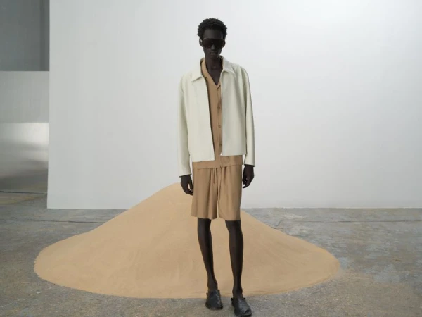 A slender man in a cream jacket and matching sand-colored shirt and shorts poses in front of a small sand dune in studio lighting.
