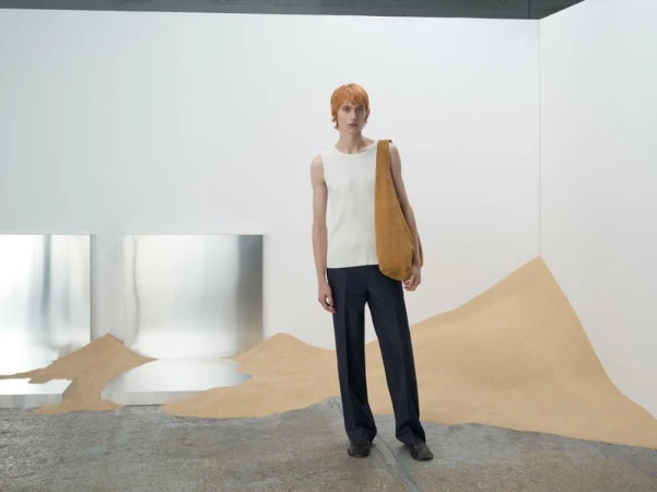 Adult male fashion model wearing a white top, navy blue pants, and carrying a tan-colored bag in a minimalist studio setting.