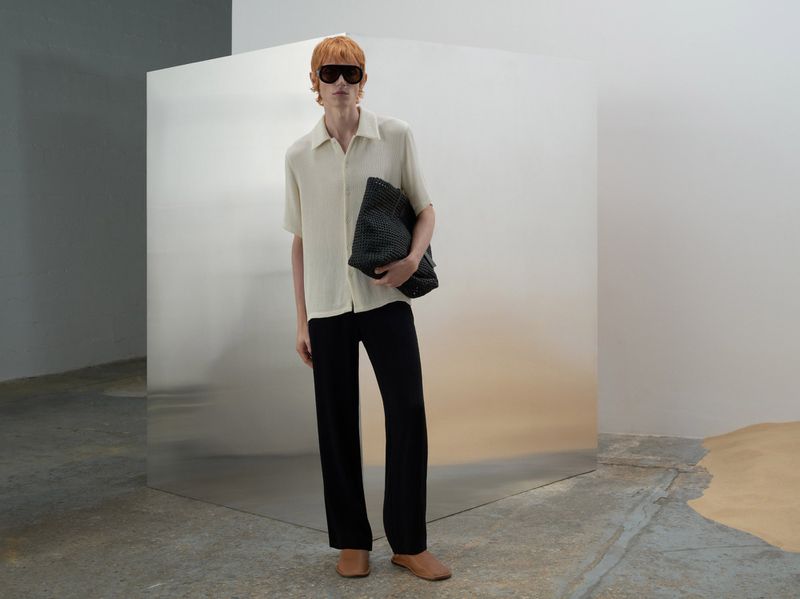 A full-length portrait of an adult man with red hair and sunglasses, wearing a neutral-toned shirt and black trousers, holding a black bag.