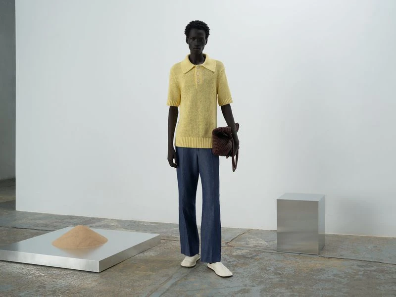Standing Black man in a butter-yellow knit polo shirt and blue jeans, holding a brown bag in a minimalist, modern indoor setting.