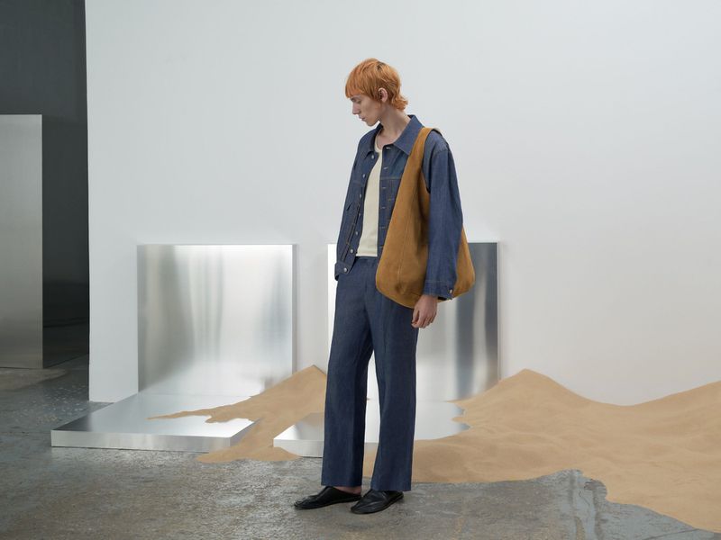A stylish adult male with red hair models a casual denim jacket, beige top, wide leg trousers, and brown bag in a studio setting.