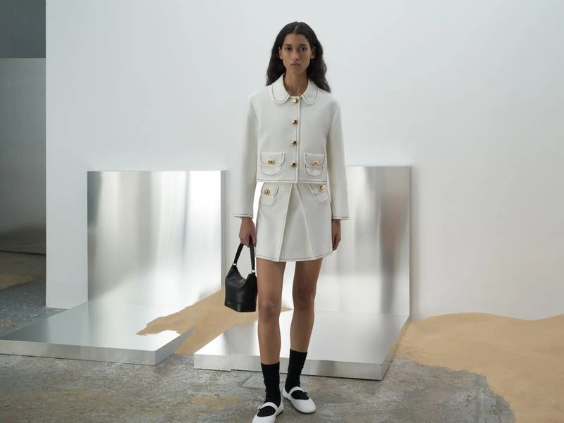 A young woman poses in a white skirt and jacket with a black purse in a neutral studio, showcasing a minimalist, elegant fashion style.