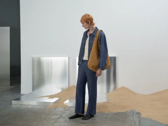 Model with orange hair wearing a blue denim jacket and matching trousers carrying a brown suede shoulder bag in a studio setting