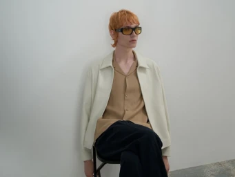 Model with orange hair and sunglasses seated wearing a light jacket, tan shirt, and dark trousers against a white wall.