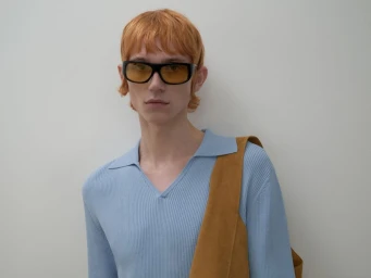 Model in amber sunglasses and a light blue polo shirt with a brown suede strap against a plain white wall