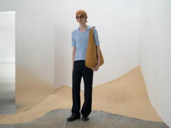 Model wearing a light blue polo shirt and dark trousers carrying a large brown shoulder bag in a white studio set with sand dunes