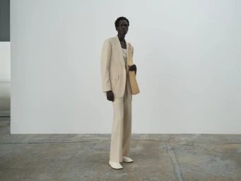 Model wearing a beige tailored suit standing against a plain white wall holding a woven shoulder bag