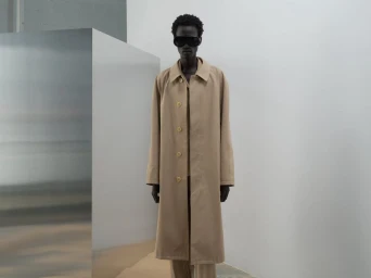 Model wearing dark sunglasses and a beige coat standing against a white studio wall with a metallic block.
