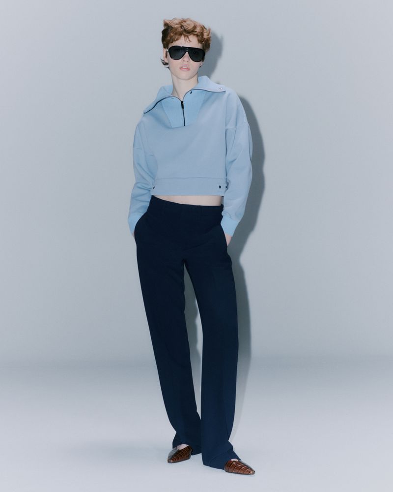 A person in a blue cropped pullover and dark trousers strikes a fashion pose in a studio with a light backdrop and wearing sunglasses.