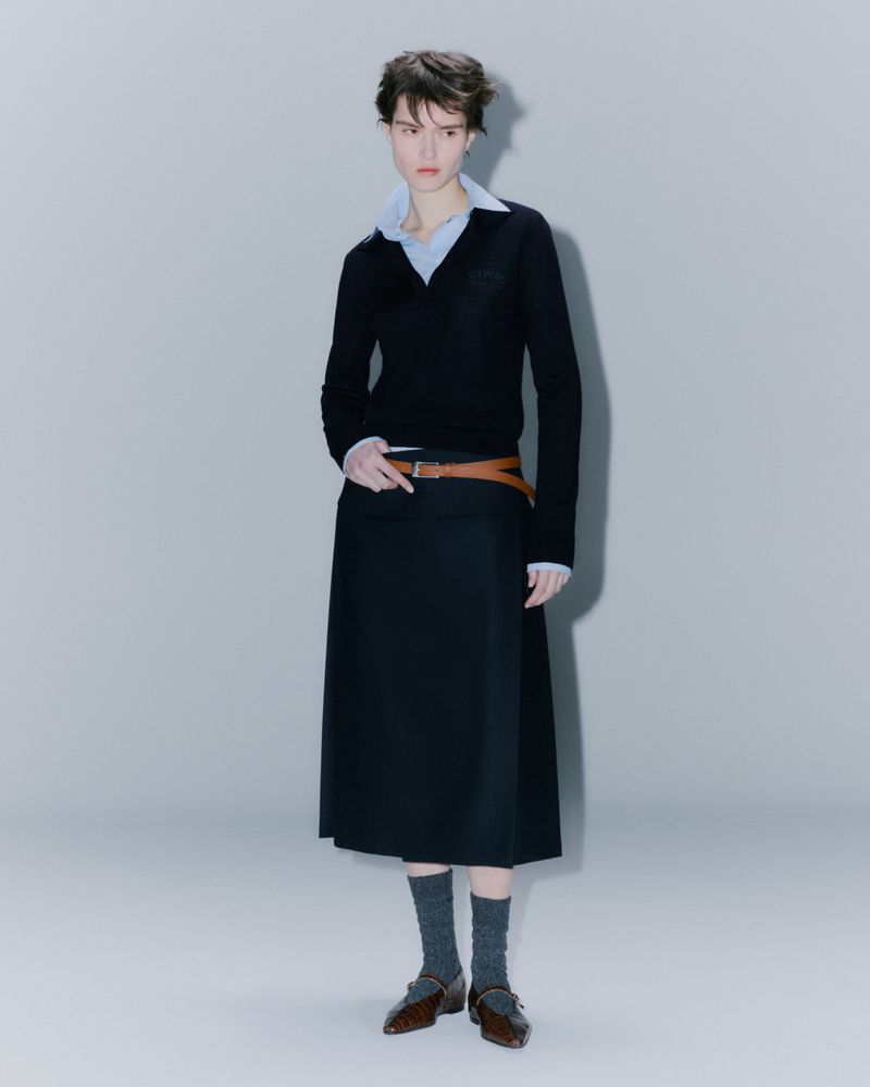 Adult woman in fashion shot. She wears a black V-neck sweater, midi skirt, brown belt, grey socks, and brown leather shoes in studio.