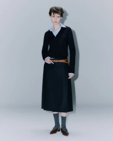Adult woman in fashion shot. She wears a black V-neck sweater, midi skirt, brown belt, grey socks, and brown leather shoes in studio.