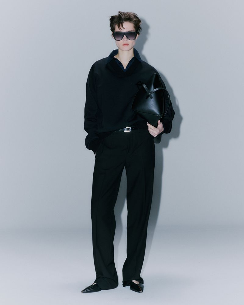 An adult female model stands in a photo studio wearing a black top and pants, sunglasses, and carrying a black handbag against a grey backdrop.