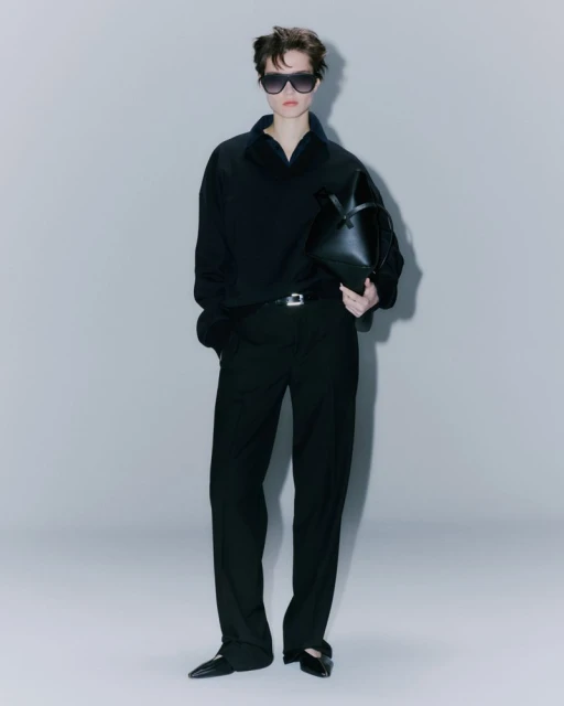An adult female model stands in a photo studio wearing a black top and pants, sunglasses, and carrying a black handbag against a grey backdrop.