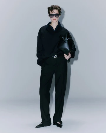 An adult female model stands in a photo studio wearing a black top and pants, sunglasses, and carrying a black handbag against a grey backdrop.