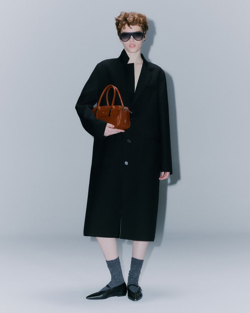 A woman in a black coat stands against a light grey background, holding a brown leather purse, complemented by dark sunglasses and flats.