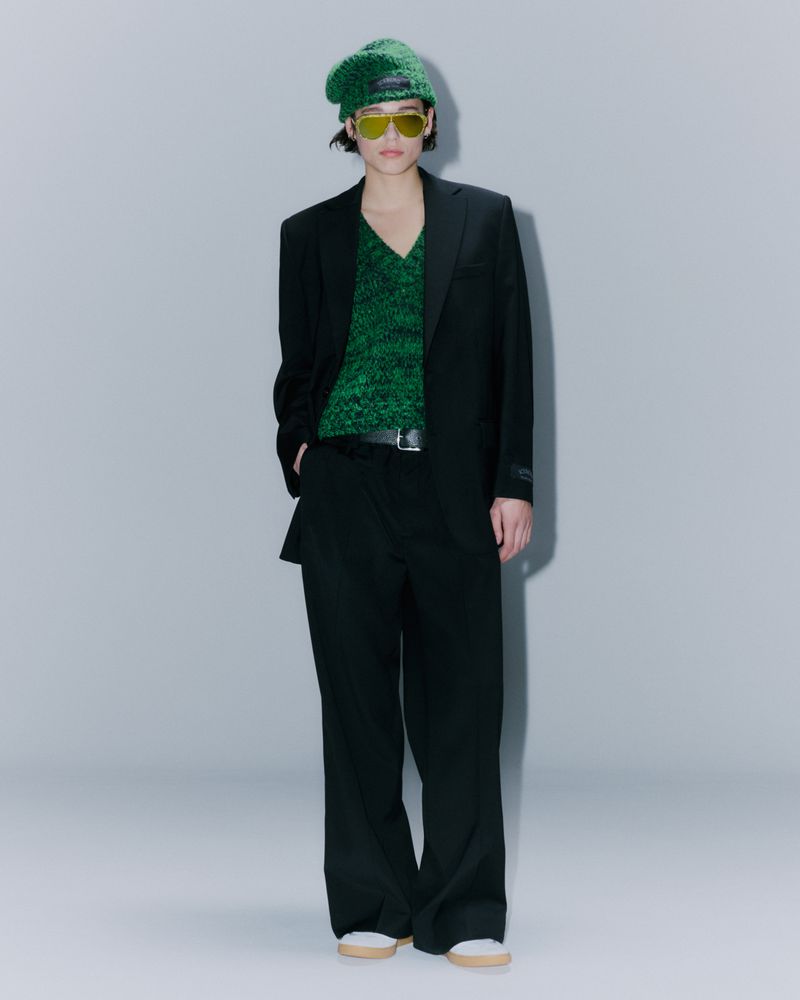 Full-length studio shot of a person wearing a black suit, green sweater, and green beret against a grey backdrop.