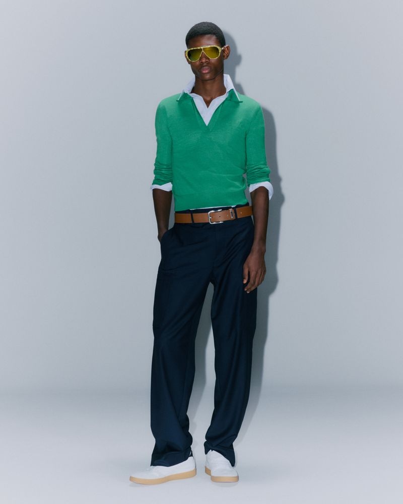 Full body studio shot of a Black male model wearing a green sweater, white collared shirt, navy pants, white shoes, and yellow sunglasses.