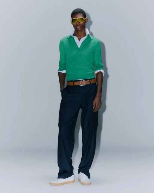 Full body studio shot of a Black male model wearing a green sweater, white collared shirt, navy pants, white shoes, and yellow sunglasses.