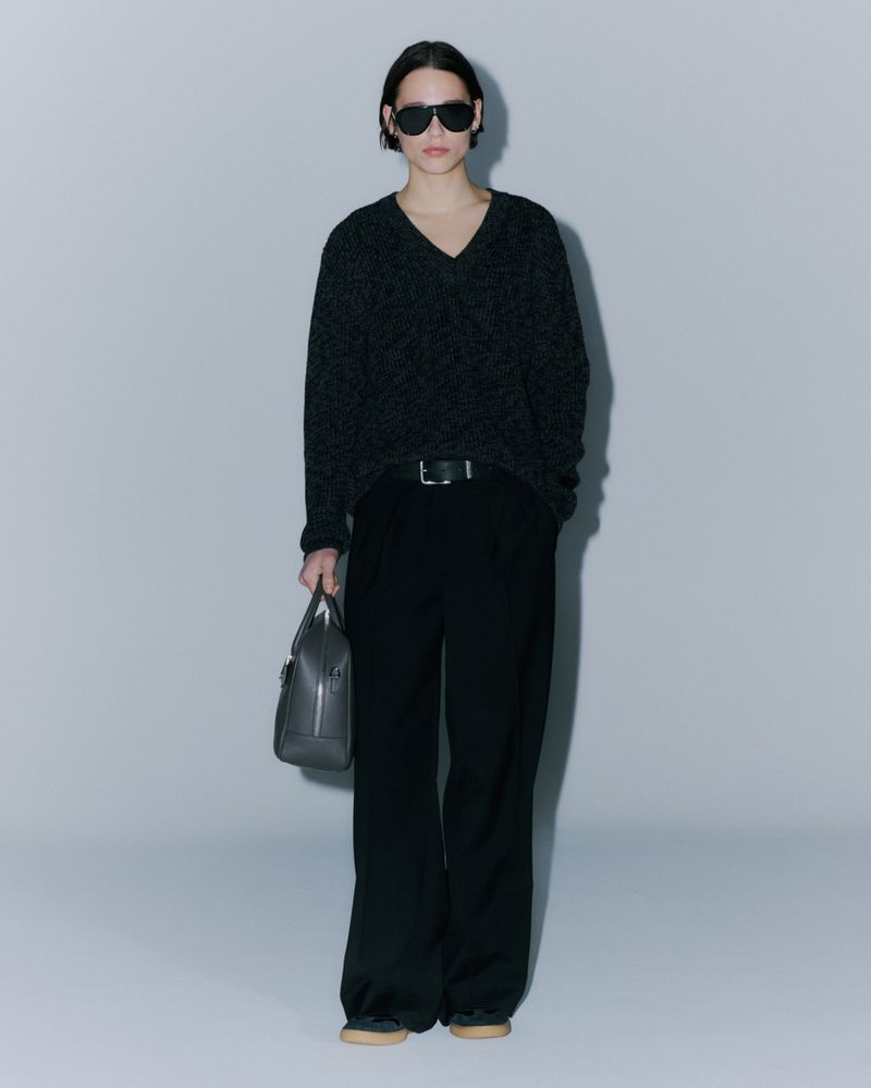 Woman in a dark v-neck sweater, black wide-leg pants, and sunglasses, holding a gray leather handbag against a light gray backdrop.