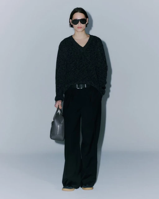 Woman in a dark v-neck sweater, black wide-leg pants, and sunglasses, holding a gray leather handbag against a light gray backdrop.