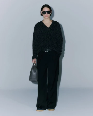 Woman in a dark v-neck sweater, black wide-leg pants, and sunglasses, holding a gray leather handbag against a light gray backdrop.