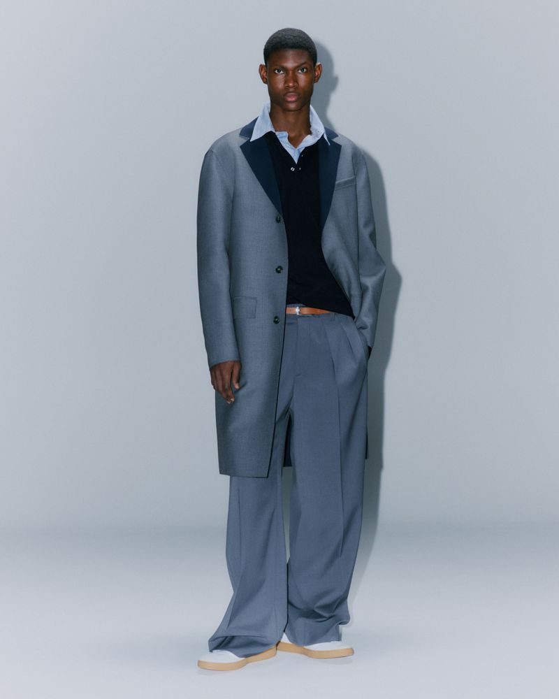 An adult male fashion model posing in a coat and trousers in a studio environment. He wears a button-down shirt and loafers.