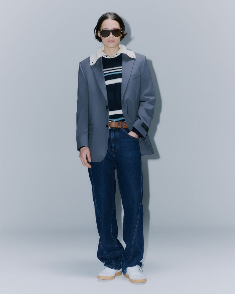 Full-length studio shot of an adult in a blue blazer with a white collar over a striped sweater, wide jeans, and sneakers, posing.