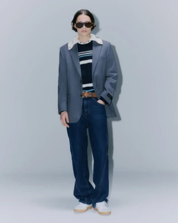 Full-length studio shot of an adult in a blue blazer with a white collar over a striped sweater, wide jeans, and sneakers, posing.