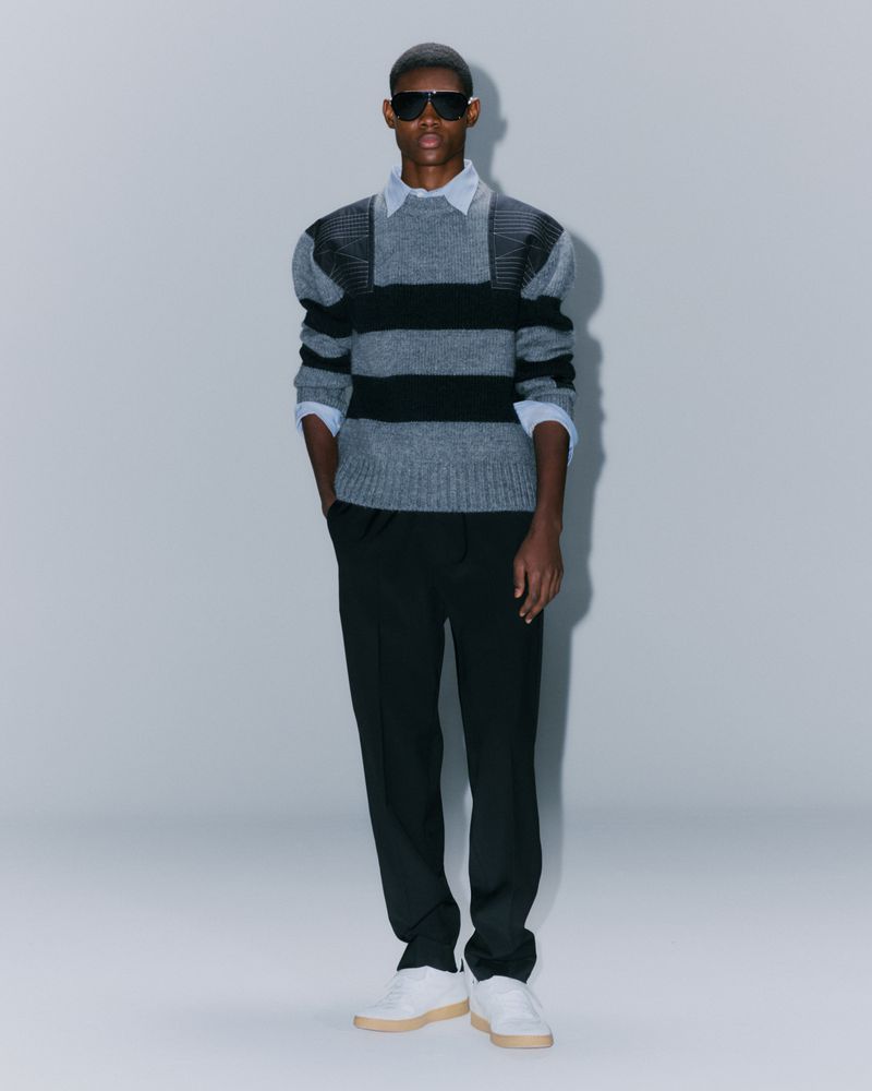 Full-length shot of a male model in sunglasses wearing a gray and black striped sweater, black pants, and white sneakers against a gray background.