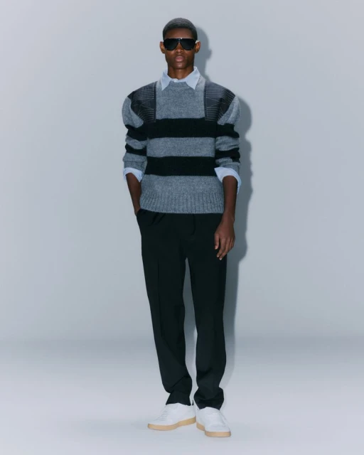 Full-length shot of a male model in sunglasses wearing a gray and black striped sweater, black pants, and white sneakers against a gray background.