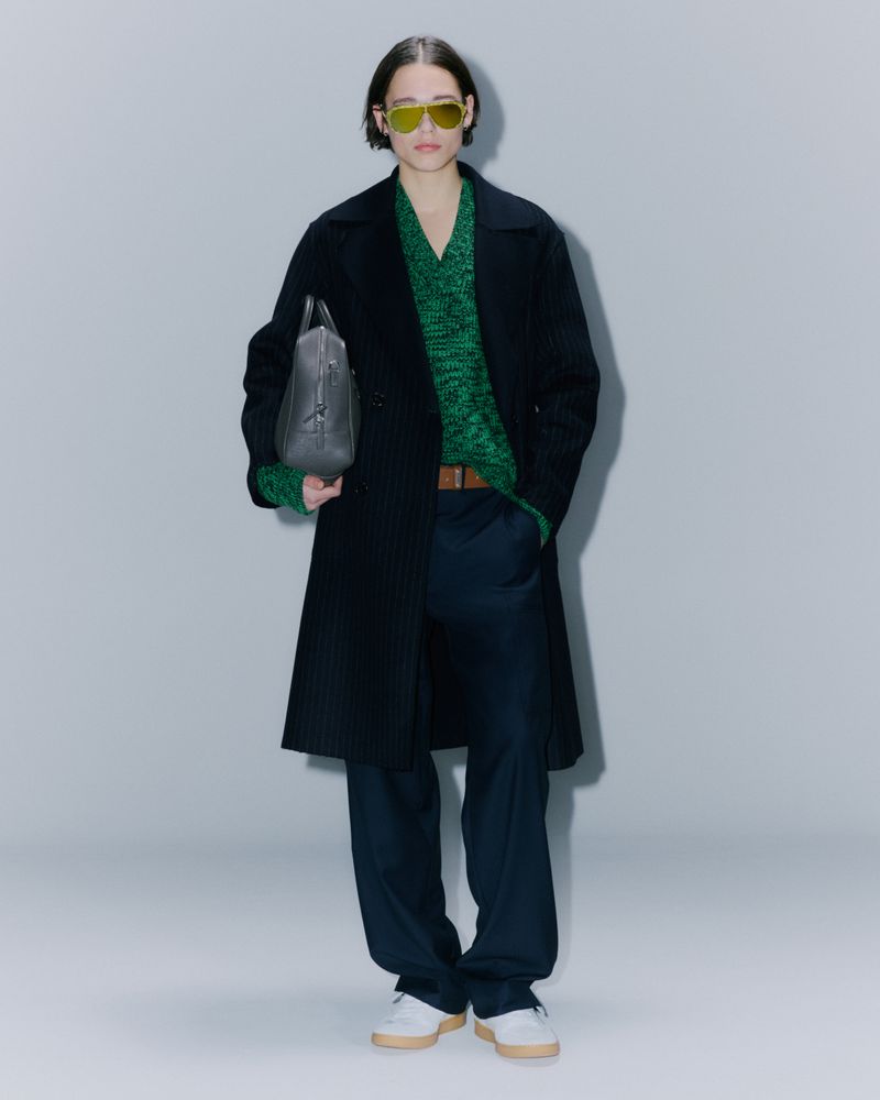 Full-length studio shot of a fashion model wearing a dark coat, green sweater, and sunglasses, holding a grey leather bag, against a grey backdrop.