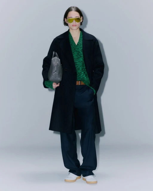 Full-length studio shot of a fashion model wearing a dark coat, green sweater, and sunglasses, holding a grey leather bag, against a grey backdrop.