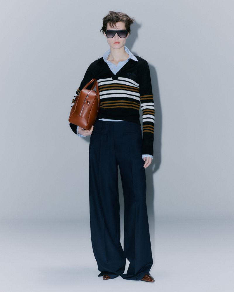 A fashion shot of a slender model in sunglasses, wearing a striped sweater, navy wide-legged trousers, and carrying a leather handbag.