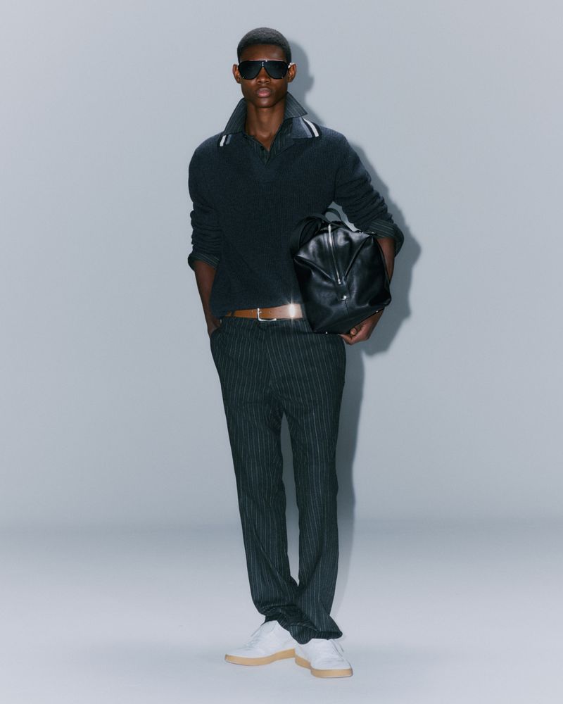Fashionable adult male in pinstripe trousers and a black collared shirt stands in a studio holding a black leather bag.