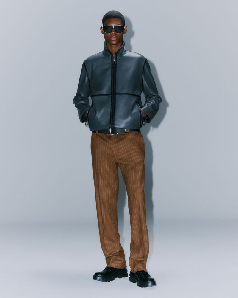 A tall adult male model poses in a full shot wearing sunglasses, a black leather jacket, brown pinstripe trousers, and black leather shoes.