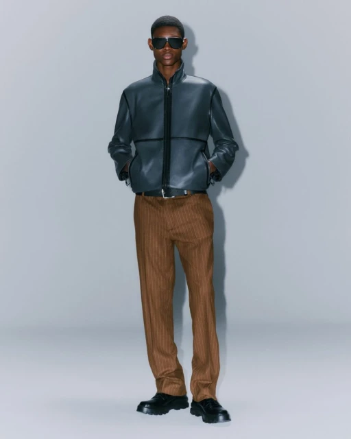 A tall adult male model poses in a full shot wearing sunglasses, a black leather jacket, brown pinstripe trousers, and black leather shoes.