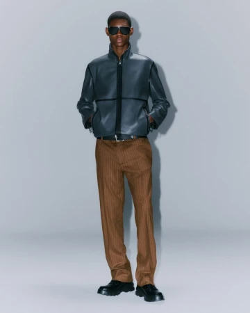 A tall adult male model poses in a full shot wearing sunglasses, a black leather jacket, brown pinstripe trousers, and black leather shoes.