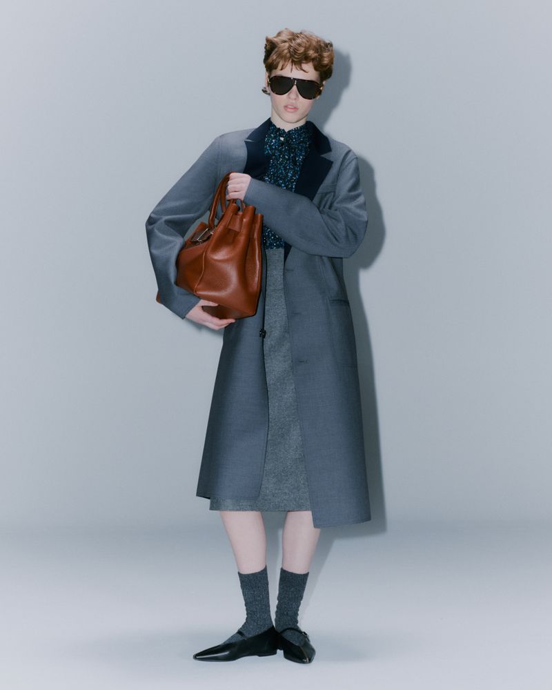 Fashionable woman in blue coat, sunglasses, and brown leather bucket bag, studio lit on neutral background, stylish and elegant.