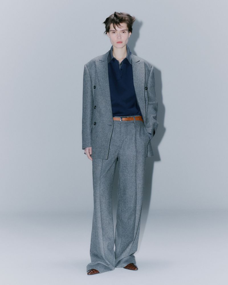 Adult fashion model poses indoors wearing a gray suit with a dark navy polo shirt and brown belt in a full-length shot.