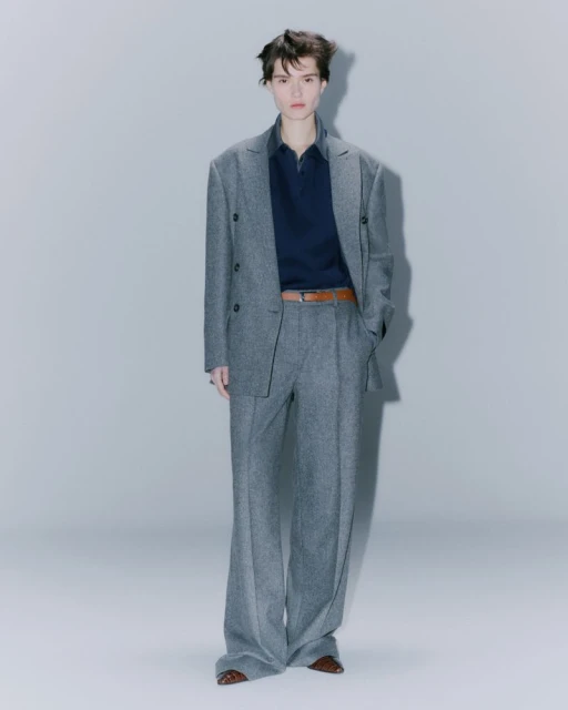 Adult fashion model poses indoors wearing a gray suit with a dark navy polo shirt and brown belt in a full-length shot.