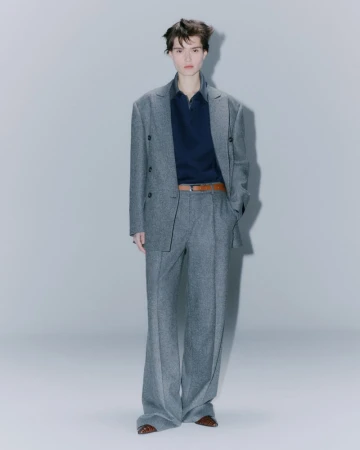 Adult fashion model poses indoors wearing a gray suit with a dark navy polo shirt and brown belt in a full-length shot.