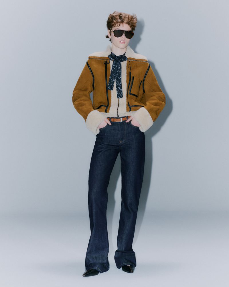 Adult female poses in a shearling jacket with dark denim jeans, a scarf, and sunglasses in a neutral studio environment.