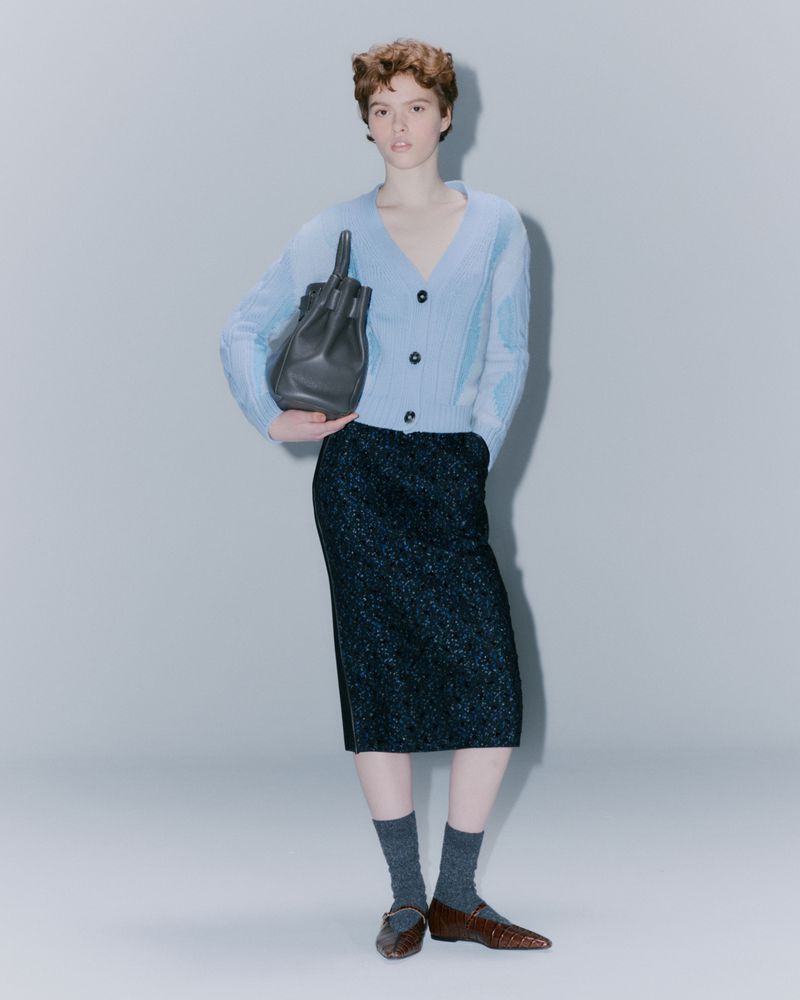 A stylish woman in a light blue cardigan and a shimmering dark skirt, holding a black handbag in a minimalist studio setting.