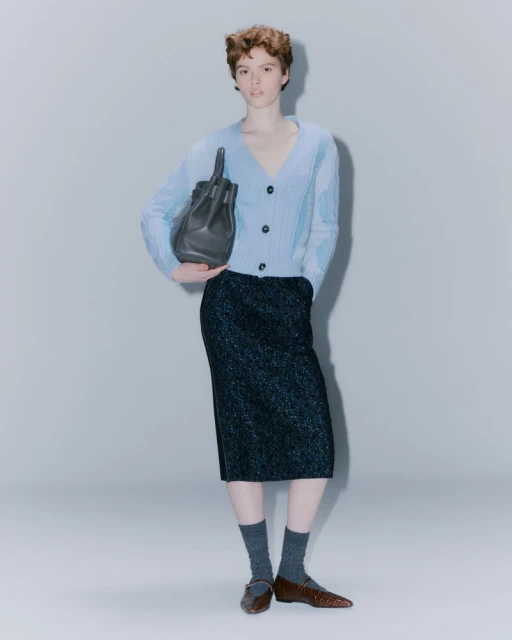 A stylish woman in a light blue cardigan and a shimmering dark skirt, holding a black handbag in a minimalist studio setting.
