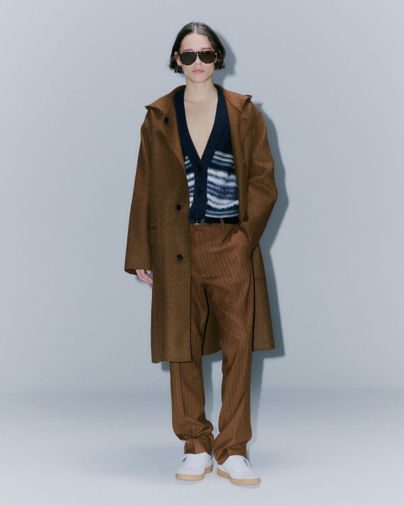 Fashionable person in a brown coat, pinstripe pants, sunglasses, and white sneakers in a full shot against a light background.